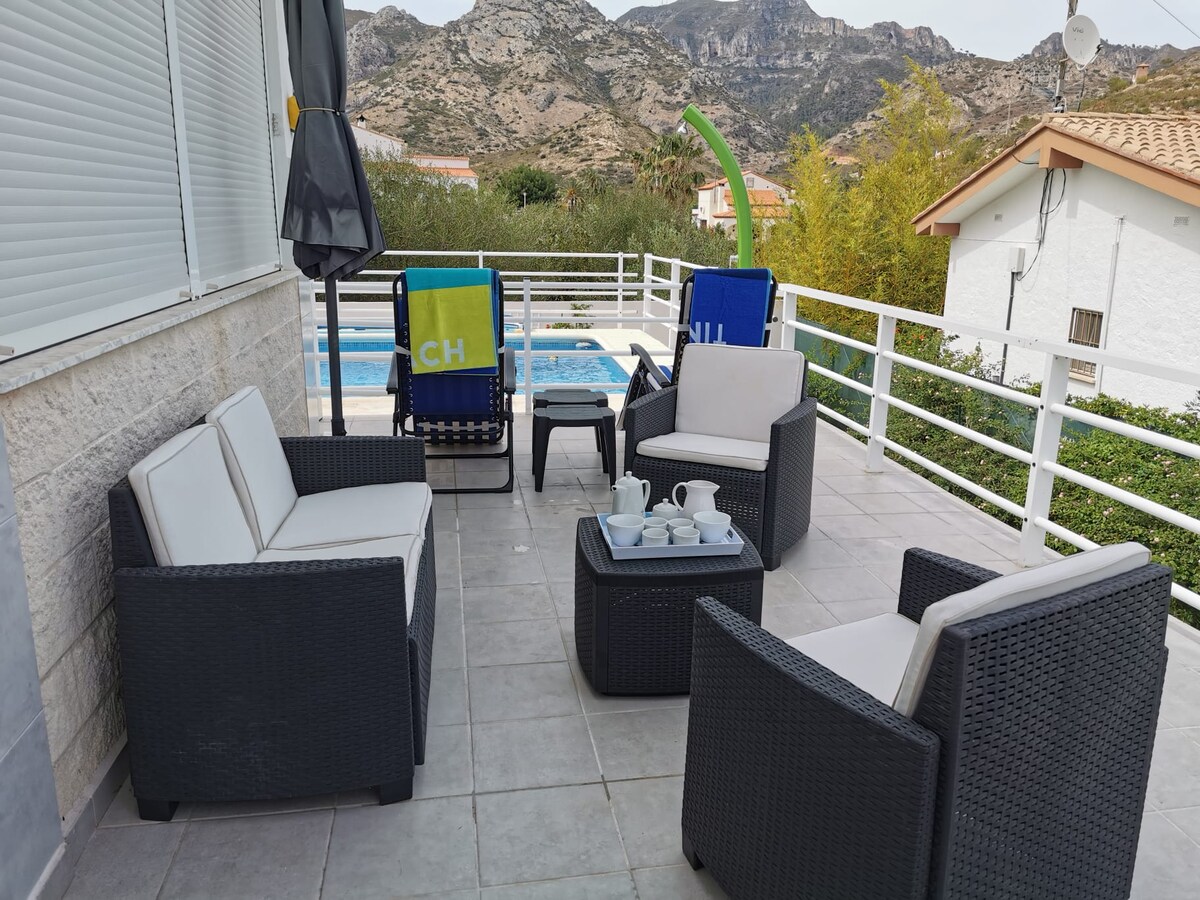 An outdoor seating area features a set of four cushioned chairs arranged around a low table. A nearby swimming pool can be seen in the background, flanked by grass and trees. The surrounding mountains provide a scenic backdrop, enhancing the peaceful ambiance.