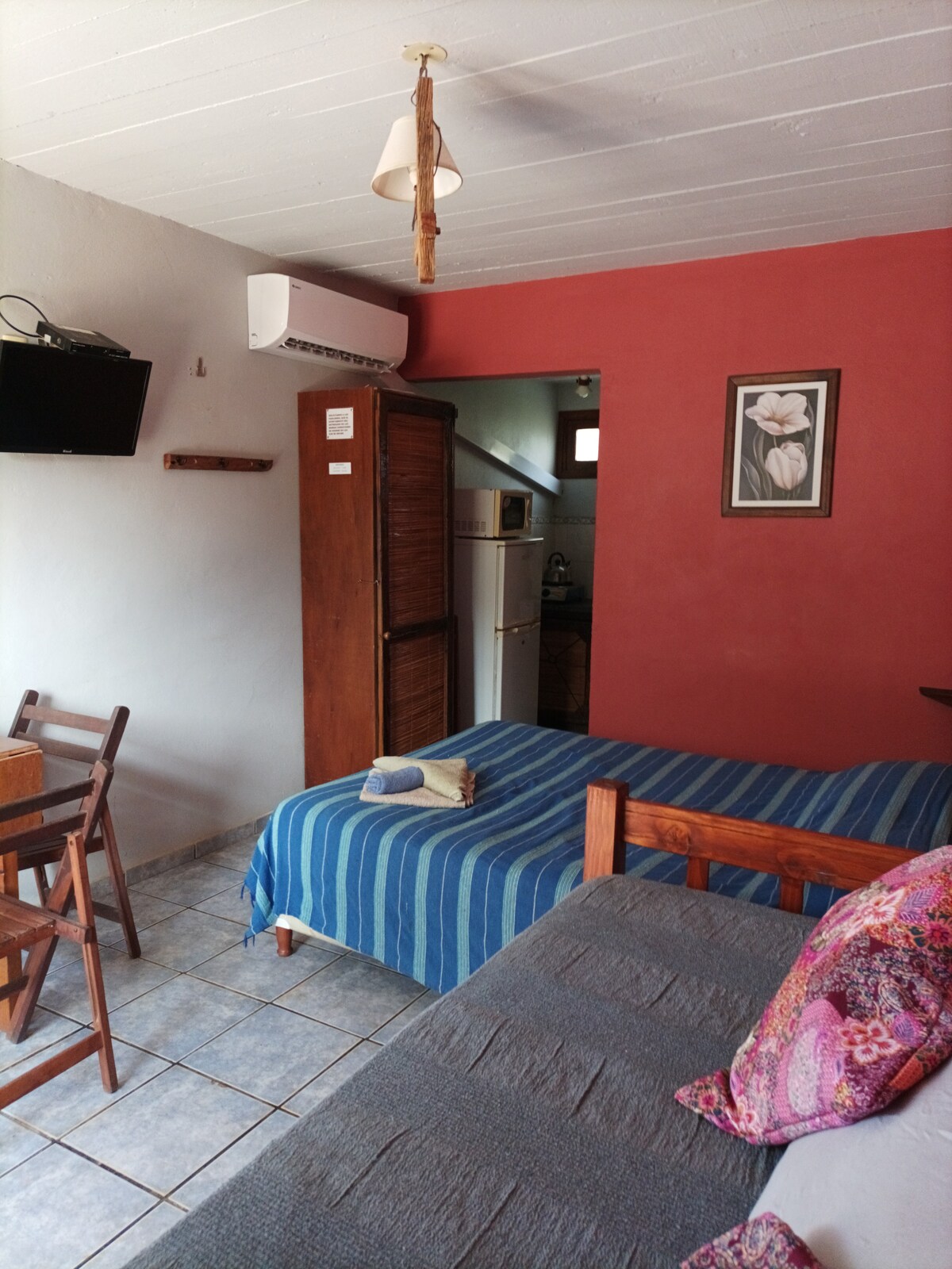 A compact sleeping space features a spacious bed covered with a blue and white striped blanket. A light grey couch is positioned nearby. A wooden table and chairs are arranged against the wall, with a flat-screen TV mounted above. A kitchen area is visible in the background.