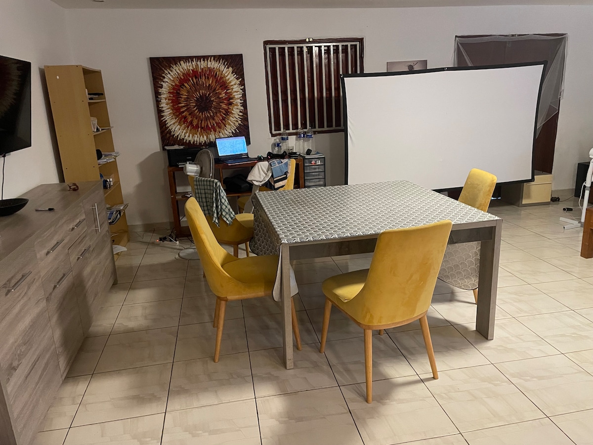 An inviting dining area is presented, featuring a rectangular table surrounded by four yellow chairs. A wall-mounted television and bookshelf are visible, along with a large screen at the back. Sunflower artwork adds a vibrant touch to the walls.