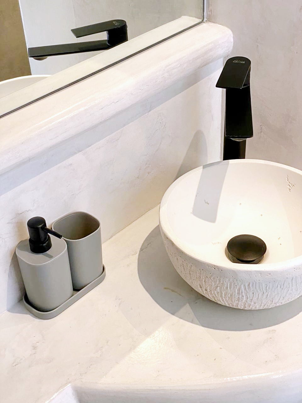 A modern bathroom sink is displayed, featuring a textured white bowl and a sleek, black faucet. Nearby, a set of gray containers is arranged neatly, holding liquid soap and a toothbrush holder. The countertop has a smooth, light-colored surface, complementing the serene atmosphere.