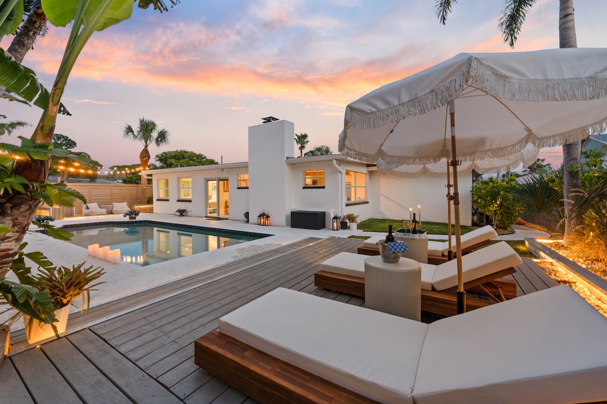 An outdoor space features a stylish lounge area with two chaise lounges placed on a wooden deck. A large umbrella offers shade over a table. The backdrop includes a resort-style pool, surrounded by palm trees, as a sunset paints the sky in soft hues.