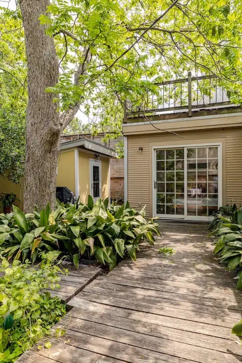 A wooden pathway leads through lush greenery towards a set of glass patio doors. A tree provides shade, while a cozy balcony above offers additional outdoor space. The surrounding plants create a serene and inviting entrance to the tranquil retreat.