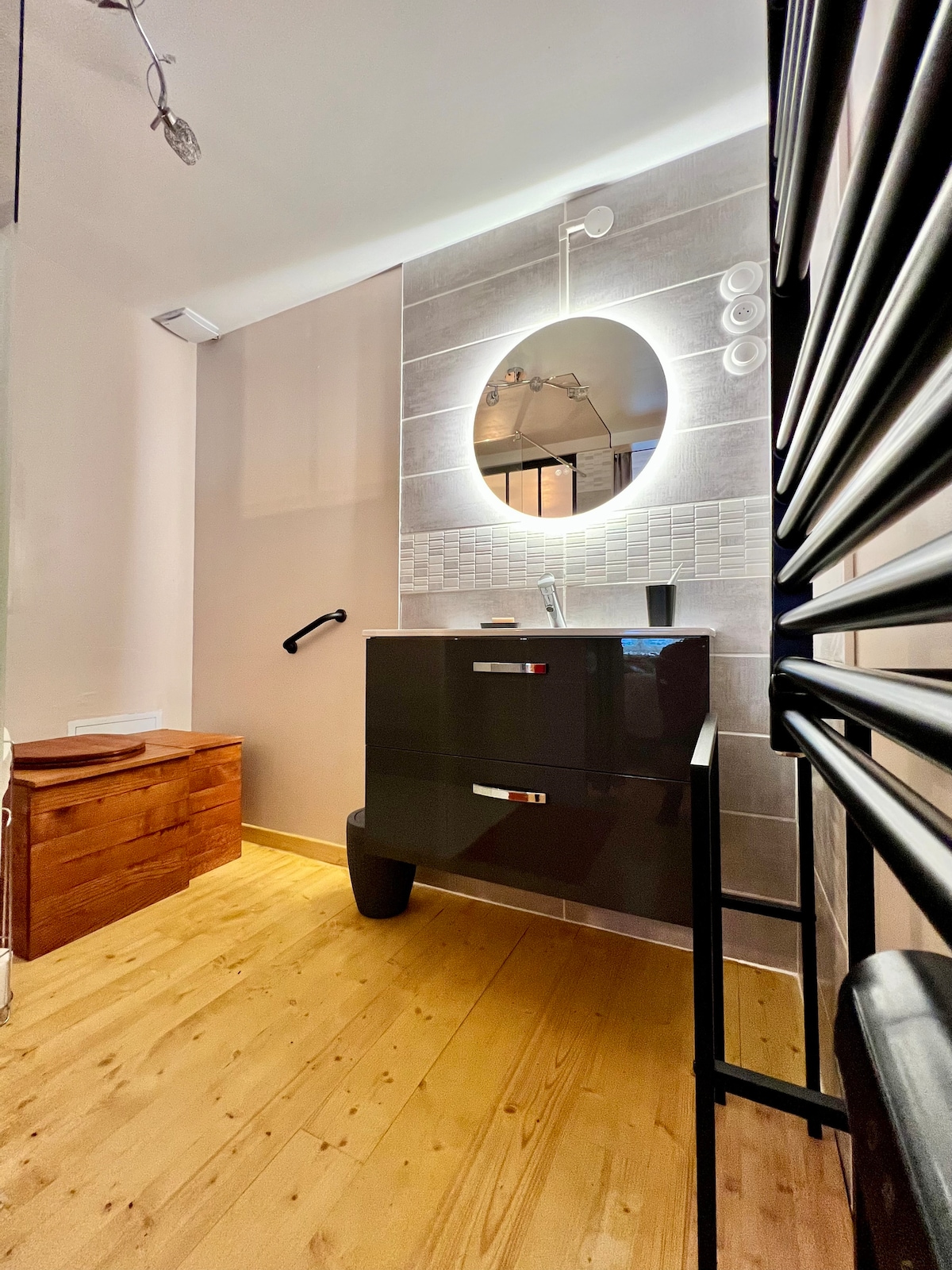 A modern bathroom is presented, featuring a sleek vanity with a round mirror illuminated by soft lighting. A wooden storage unit is positioned to the left, and a towel radiator occupies the right side. The space is finished with neutral wall tiles and warm wooden flooring.