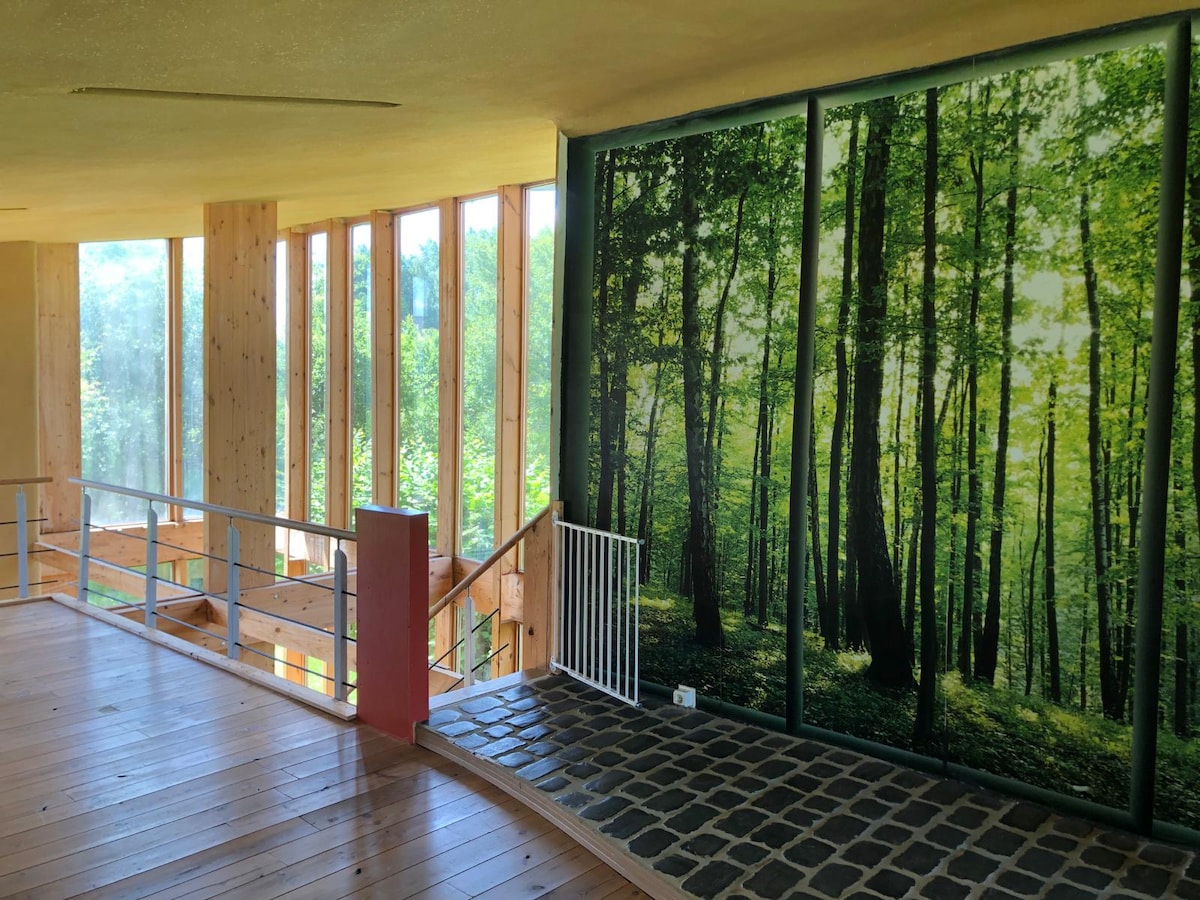 A spacious interior area is highlighted by large windows allowing natural light to fill the space. A forest-themed mural adds an element of nature to the surrounding wooden walls. A railing lines the open walkway, providing safety and an unobstructed view.