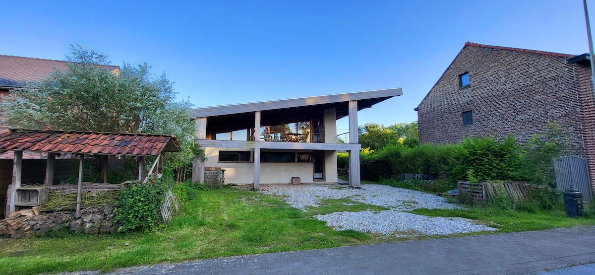 The exterior of a modern ecologically designed wooden house is visible, featuring a spacious covered terrace and large windows. Surrounding greenery adds to the natural setting. Nearby, an old structure with a rustic roof appears, alongside a gravel pathway leading to the entrance.