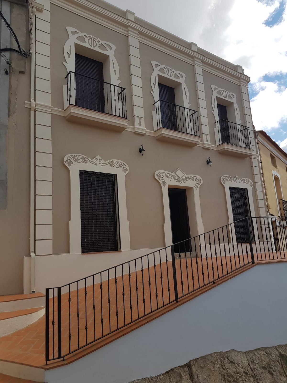 A charming building features elegant architectural details, including decorative moldings and wrought iron balconies. The sloped pathway leads to the entrance, while large, closed windows offer a sense of privacy. The facade is painted in a neutral tone, contributing to a welcoming exterior.