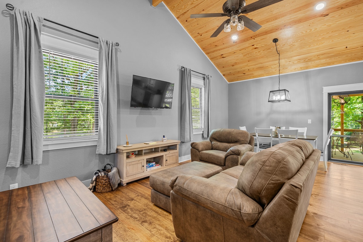 A spacious living area is highlighted by a light wood ceiling and large windows that allow natural light to fill the room. A comfortable brown sectional sofa is positioned near a flat-screen TV. A dining table is seen in the background, inviting gatherings.