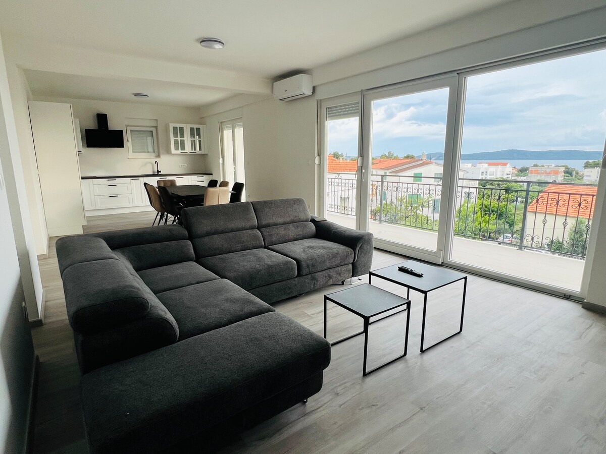 A spacious living area is highlighted by a dark gray sectional sofa, positioned across from two black coffee tables. Large windows provide natural light and reveal views of the surrounding area and sea beyond. A dining table for four is visible in the background.