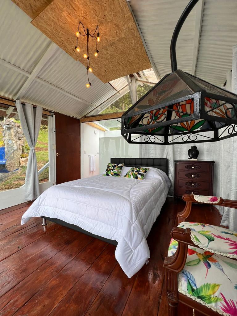 A bright sleeping area features a spacious bed adorned with a crisp white duvet. A bedside table holds a decorative lamp, while a vintage chair showcases floral upholstery. Sheer curtains allow natural light to filter in, enhancing the airy feel of the room.