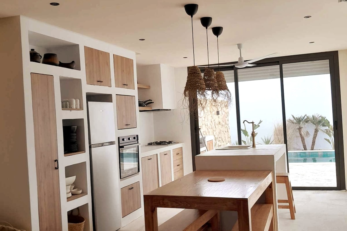 A modern kitchen features light wood cabinetry and open shelving, showcasing various dishware. A large island serves as a dining space, accompanied by stools. Ample natural light filters through expansive glass doors, revealing views of the pool and surrounding palm trees.