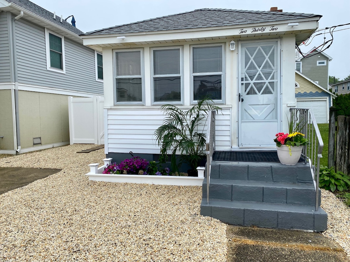 A charming cottage exterior is displayed, featuring a white siding facade with a welcoming front entrance. Steps lead to a covered porch adorned with potted flowers. Surrounding gravel enhances the clean and tidy appearance, complemented by small shrubs in the garden beds.
