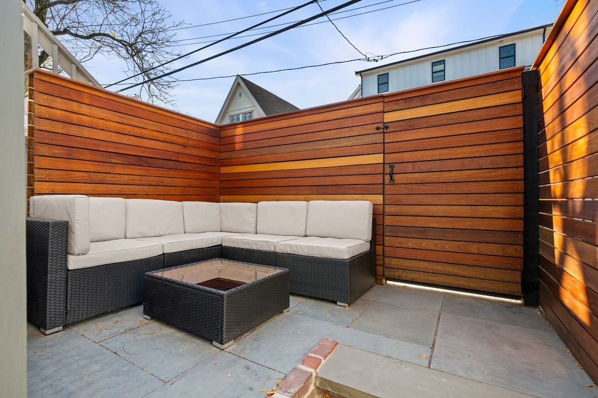 An outdoor seating area features a corner sectional sofa with light upholstery, surrounding a low fire pit on a stone patio. The space is enclosed by wooden fencing, providing privacy while allowing natural light to filter through.