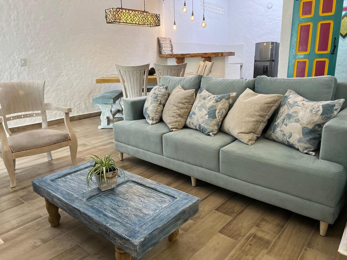 A comfortable living space is shown, featuring a soft, light blue sofa adorned with patterned cushions. A rustic wooden coffee table is positioned in front, and a potted plant adds a natural element. Bright lighting is provided by pendant fixtures above.