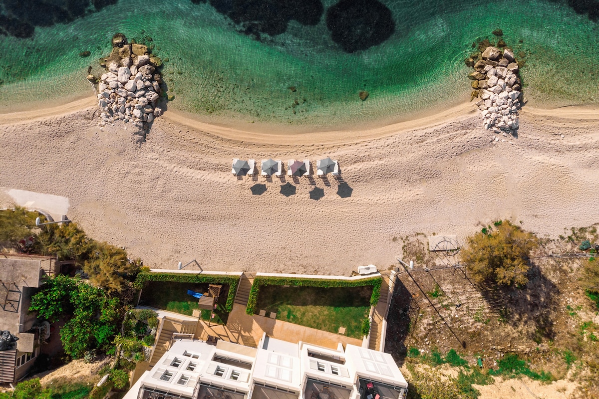 An aerial view captures the serene beach with gentle waves lapping at a sandy shoreline. Sunloungers are arranged for relaxation under minimalist shades. The water is a clear gradient of greens and blues, while natural rock formations frame the beach, enhancing the tranquil environment.