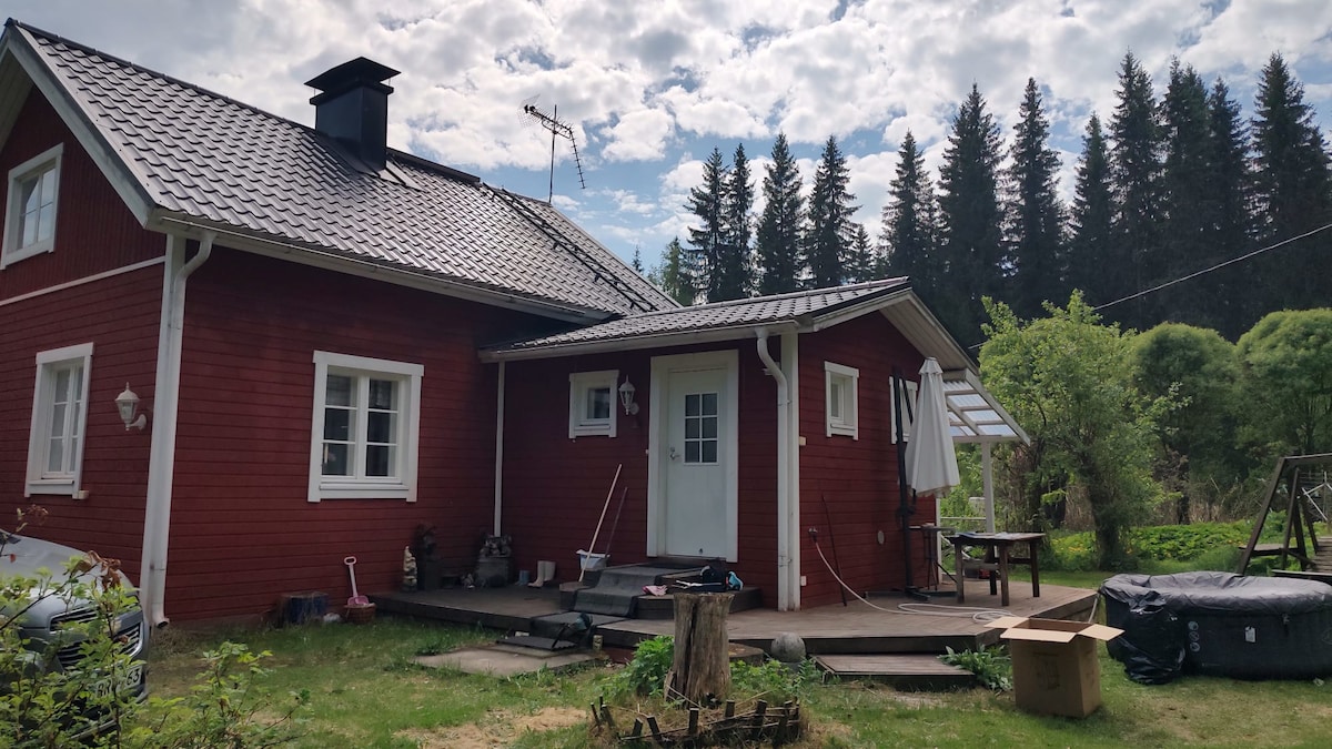 A red wooden cabin with a sloped roof is set within a natural landscape, surrounded by trees. A spacious wooden deck is visible, featuring a table and chairs. Sunlight filters through the clouds, highlighting the serene outdoor environment.