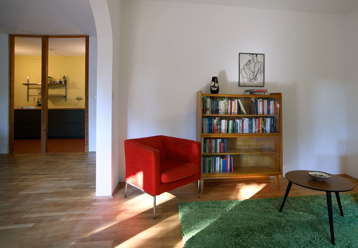 A bright living area is highlighted by a vibrant red armchair and a wooden bookshelf filled with books. Light filters in, illuminating a green rug and a small coffee table. The open kitchen can be seen in the background, providing a sense of spaciousness.