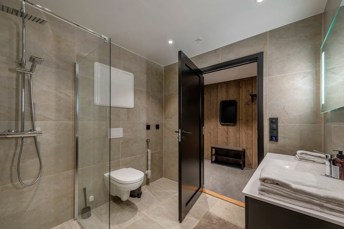 A modern bathroom is presented with a glass shower enclosure and a wall-mounted toilet. Natural stone tiles cover the floor and walls, while a sleek vanity with neatly arranged towels and toiletries enhances the overall design. A door leads into an adjoining area.