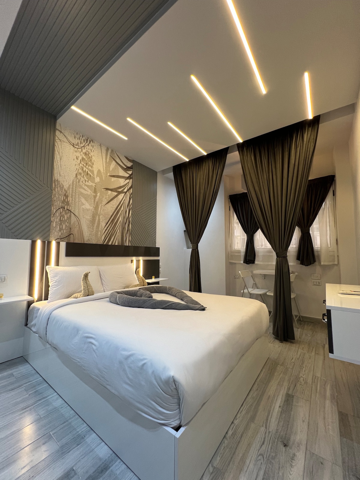 A modern bedroom features a spacious bed with neutral bedding, accented by decorative pillows. Floor-to-ceiling curtains frame a window, allowing natural light to softly illuminate the room. Sleek, contemporary design elements are showcased through distinct wall patterns and subtle lighting.