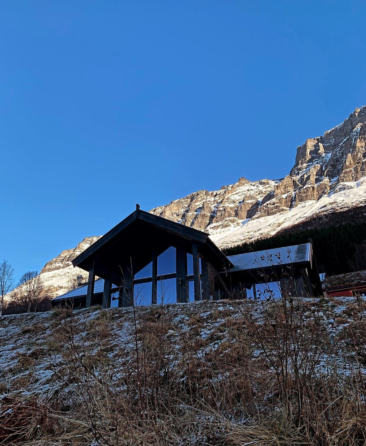 A modern cabin is positioned against a backdrop of snow-capped mountains, with large glass windows reflecting the blue sky. The surrounding area is characterized by sparse vegetation, hinting at natural beauty and tranquility.