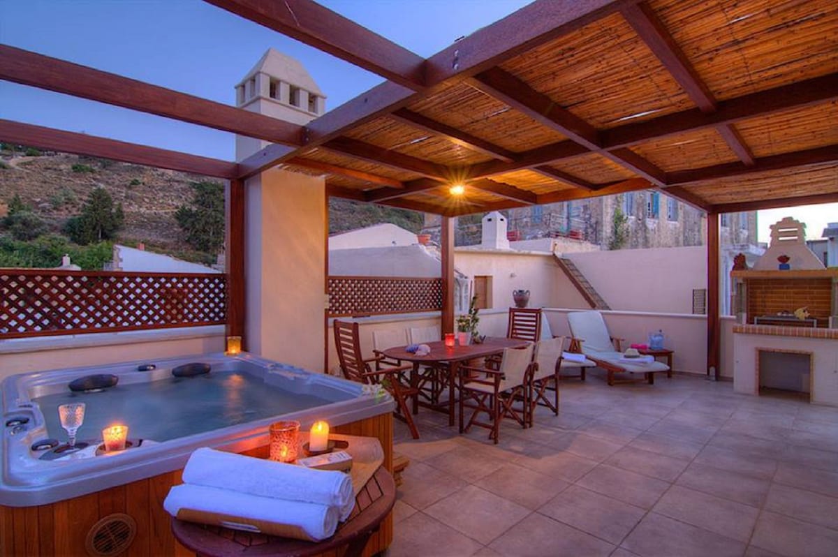 A private outdoor area features a Jacuzzi surrounded by wooden decking. Comfortable lounge chairs and a dining table are arranged for relaxation. Soft lighting creates a serene ambiance, while a mountainous backdrop adds to the tranquil setting.
