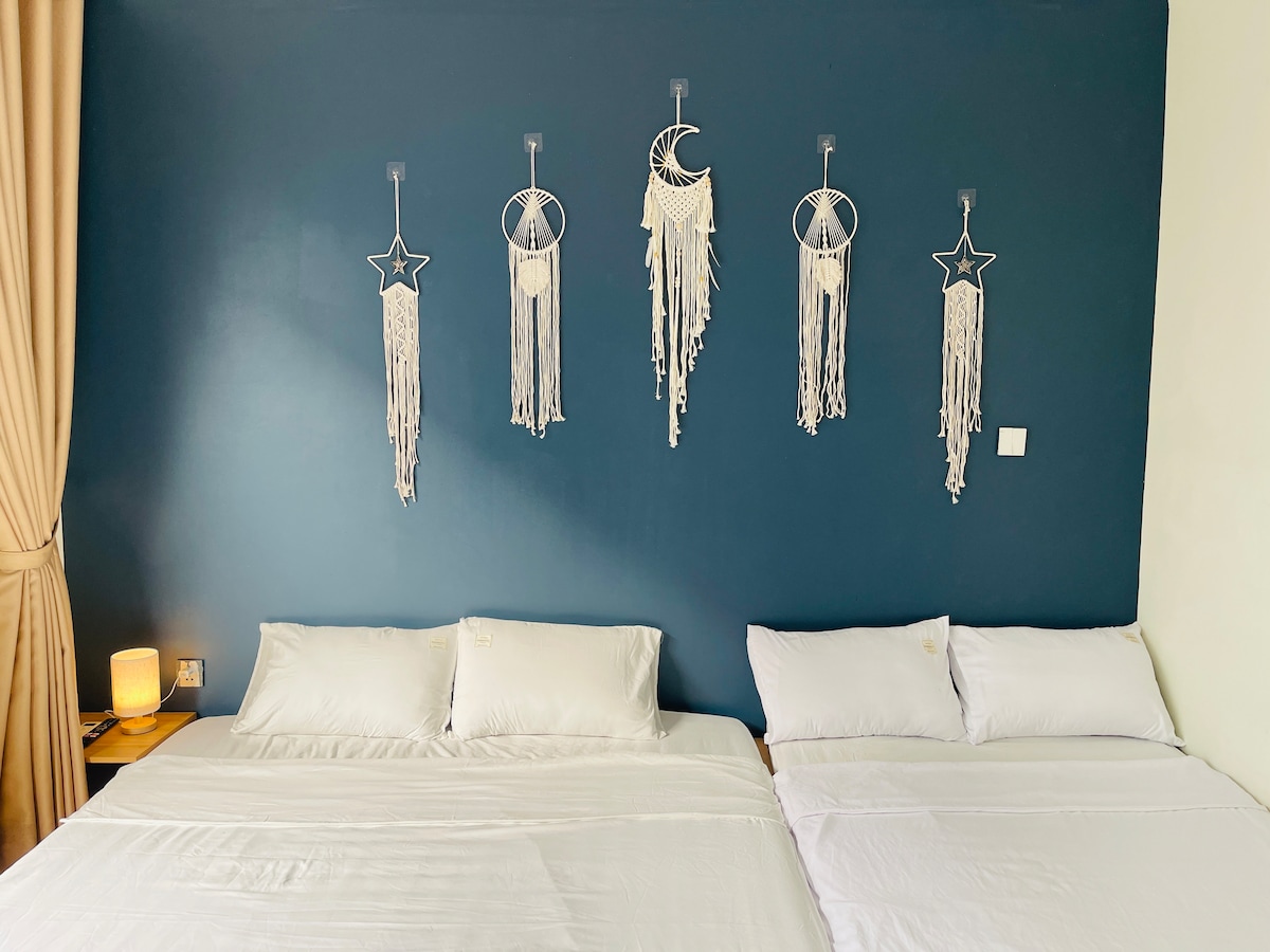 A serene sleeping area features two spacious beds, each dressed in crisp white linens. The deep blue wall is adorned with decorative macramé hangings, adding an artistic touch. Soft lighting is provided by a bedside lamp, contributing to the overall ambiance.