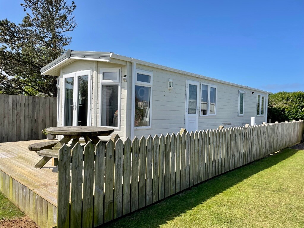 A single-story static caravan is positioned within a fenced area, featuring a deck with outdoor seating. Large windows allow natural light to fill the interior, while a tree and grassy lawn provide a pleasant outdoor setting.