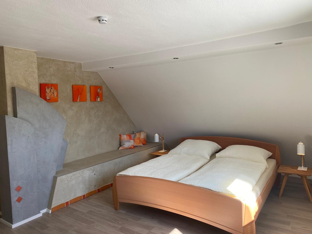 A spacious sleeping area showcases a large wooden bed with white bedding. A cozy seating nook is visible along the wall, complimented by orange accents. Bright wall art adds a pop of color, while natural light streams in from a nearby window.