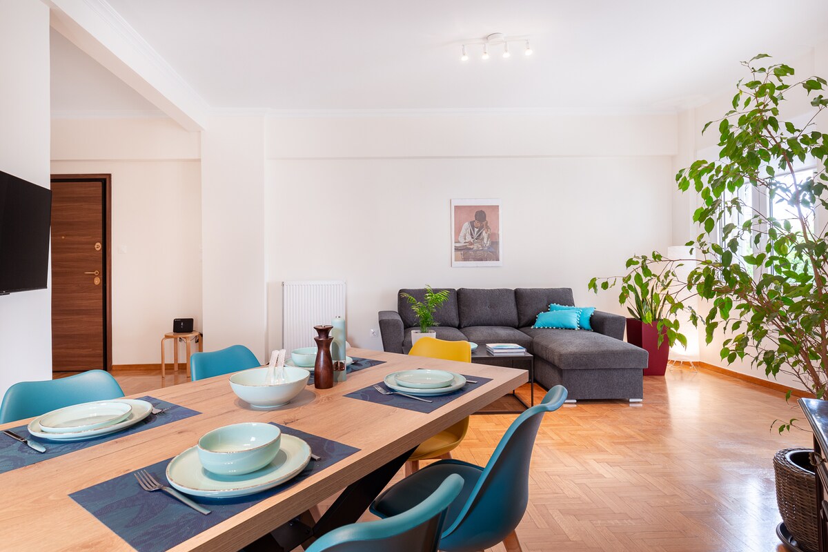 An open-plan living and dining area is presented, featuring a wooden dining table set for six. A grey sectional sofa is positioned nearby, accented with turquoise cushions. A vibrant green plant adds a touch of nature to the space, illuminated by natural light from the window.