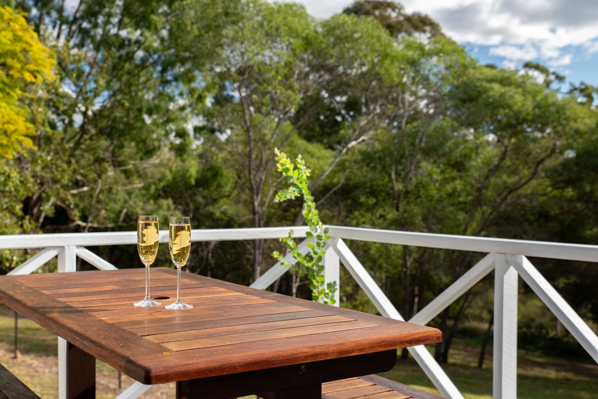A wooden table is set on a private deck, featuring two glasses of sparkling wine. Surrounding greenery offers a serene backdrop, with trees and distant hills visible under a bright sky.