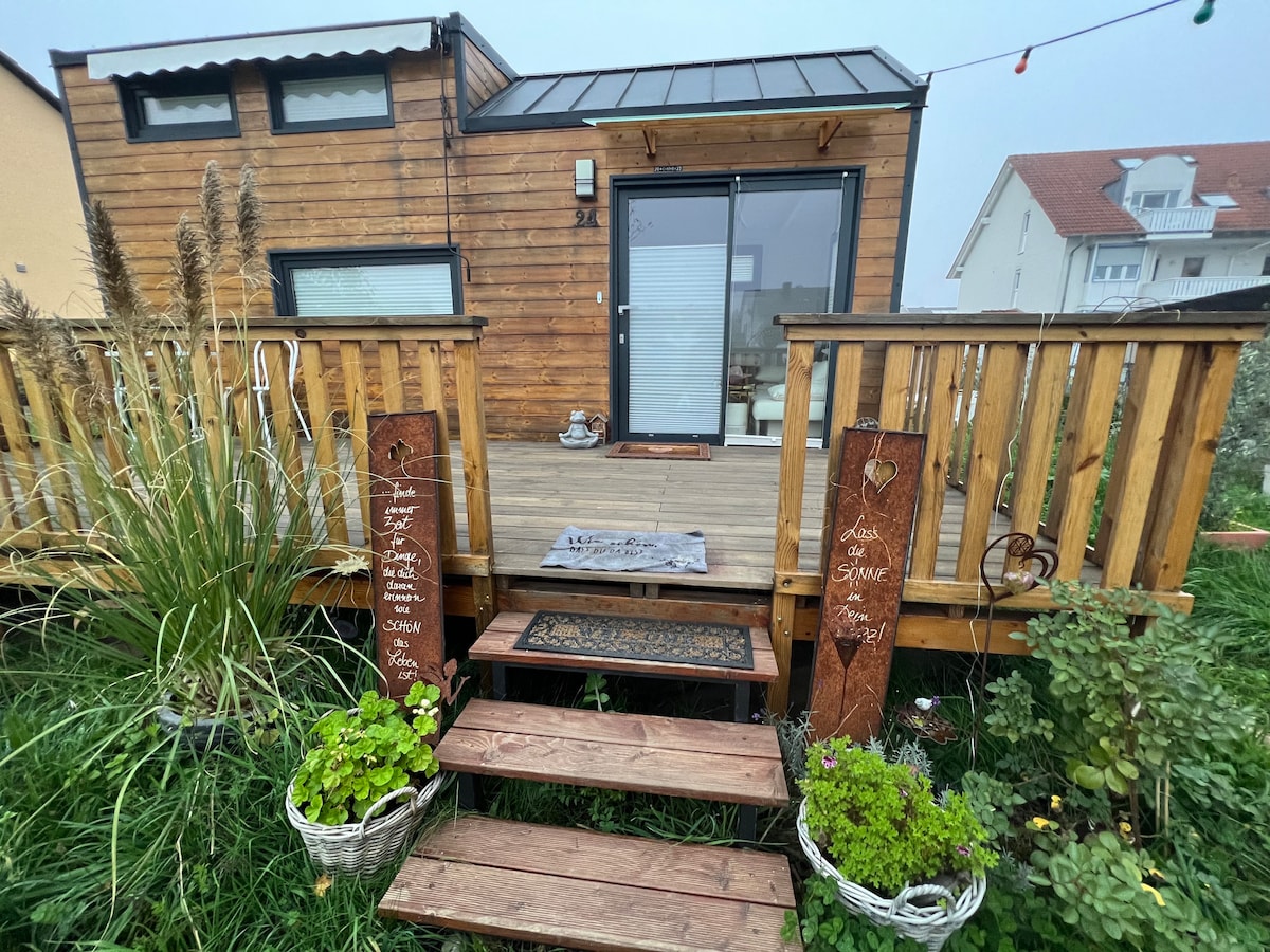 A wooden deck leads to the entrance of the tiny house, featuring large glass doors that invite natural light. Potted plants and flowering grasses adorn the entryway, enhancing the greenery. Step stones guide the way to the door, adding charm to the outdoor space.