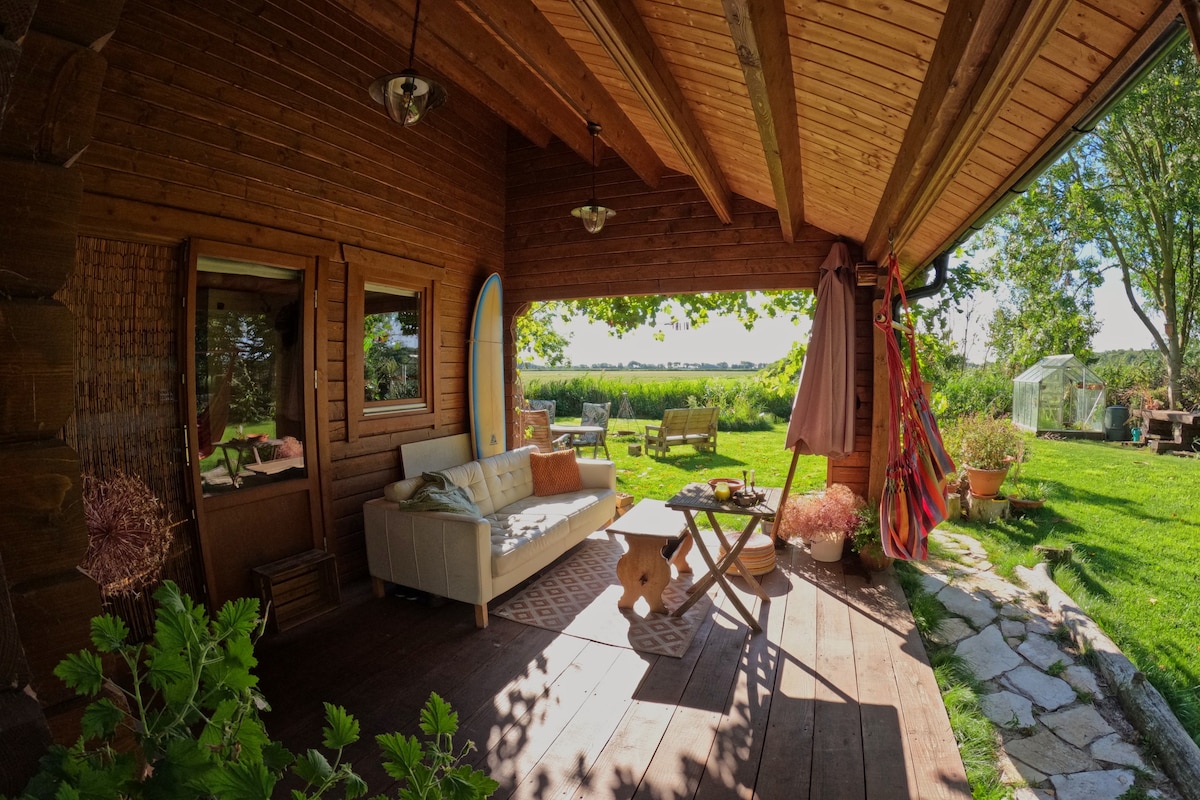 A spacious outdoor seating area is featured on a wooden deck, showcasing a comfortable sofa and a rustic wooden table. Bright sunlight filters through the open space, illuminating the lush garden and surrounding fields. A surfboard leans against the wall, and vibrant plants add a touch of greenery.