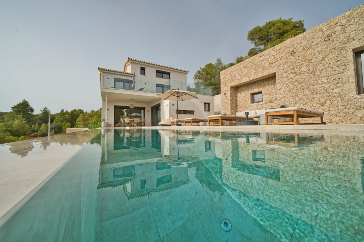 A modern villa is depicted from a low angle, emphasizing the clear water of the pool in the foreground. The villa's stone facade and expansive outdoor seating area are visible, framed by a backdrop of lush greenery and the serene architecture of the upper floors.