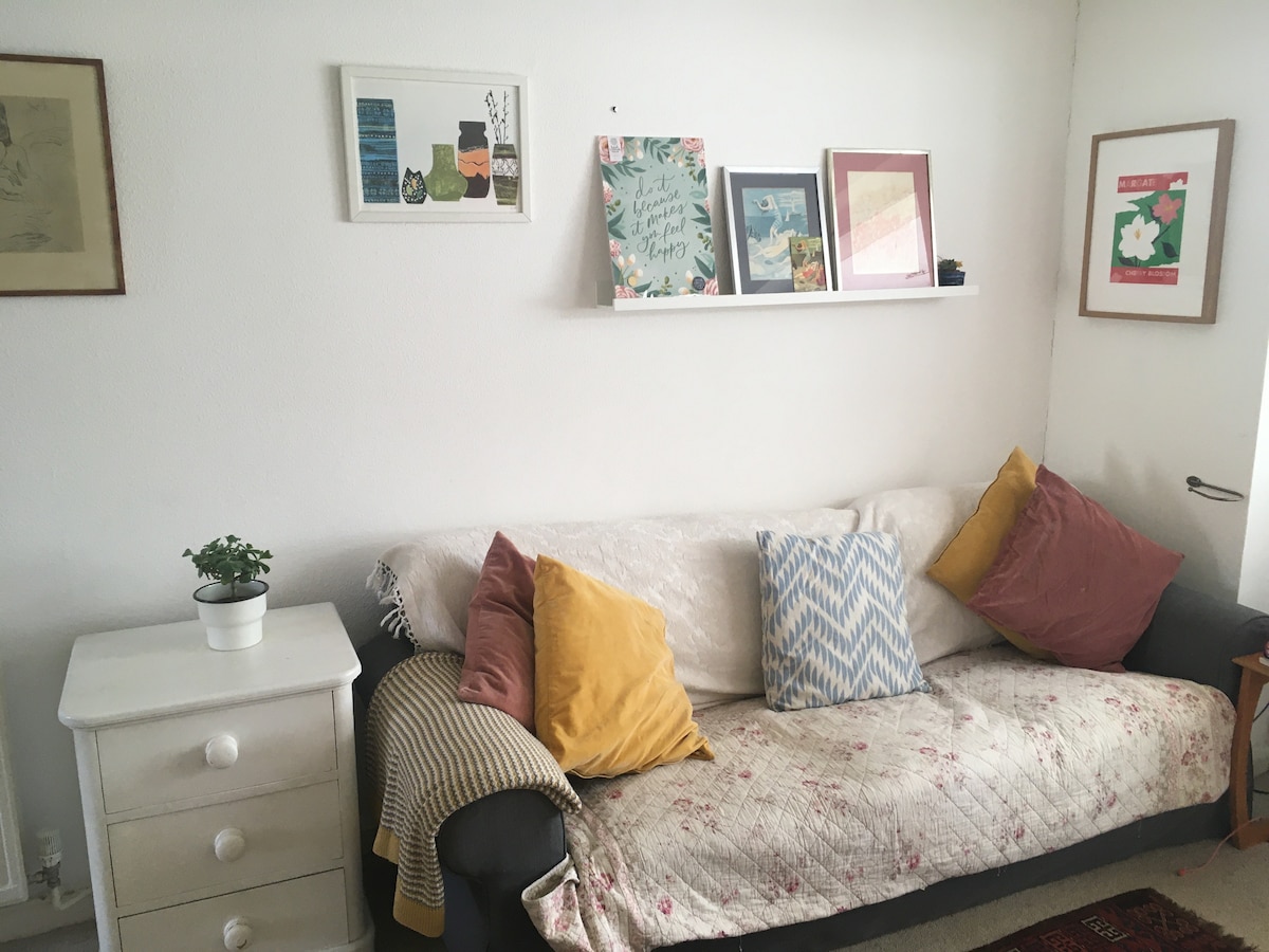 A cozy seating area features a sofa adorned with a variety of pillows. A light quilt drapes over the sofa, while a small plant sits on a nearby white nightstand. The walls display a selection of framed artwork and decorative prints.