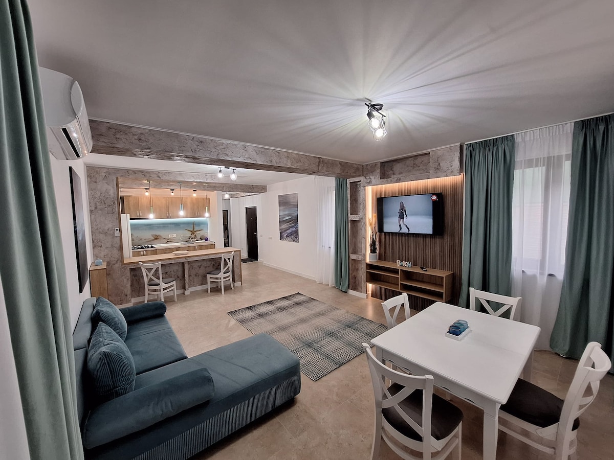 An open-plan living area is featured, incorporating a comfortable seating arrangement with a large sectional sofa. A dining table sits nearby, complemented by bright curtains. The kitchen is visible in the background, equipped with modern appliances. A television is mounted on the wall, enhancing the space.