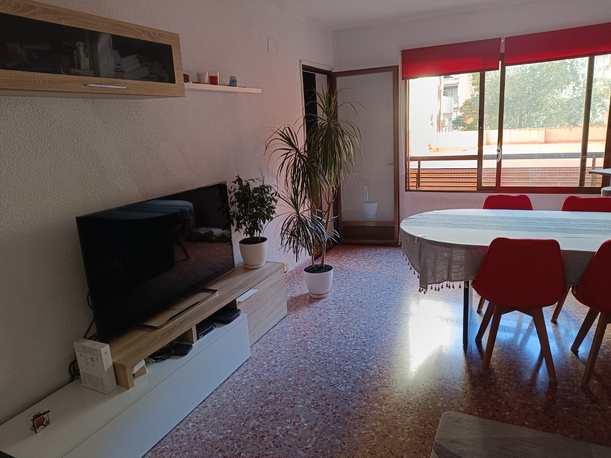 A spacious living area is featured, showcasing a large dining table surrounded by red chairs. A flat-screen TV is positioned on a light-colored entertainment unit. Natural light pours in through expansive windows, illuminating the room and highlighting a potted plant near the window.