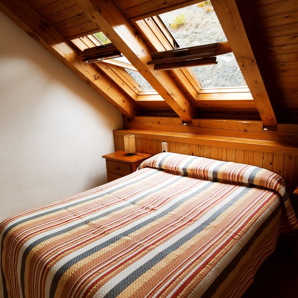 A cozy bedroom features a spacious bed dressed in a striped quilt. Natural light enters through two skylights, illuminating wooden beams and paneling. A bedside table is positioned next to the bed, enhancing the intimate and warm ambiance.