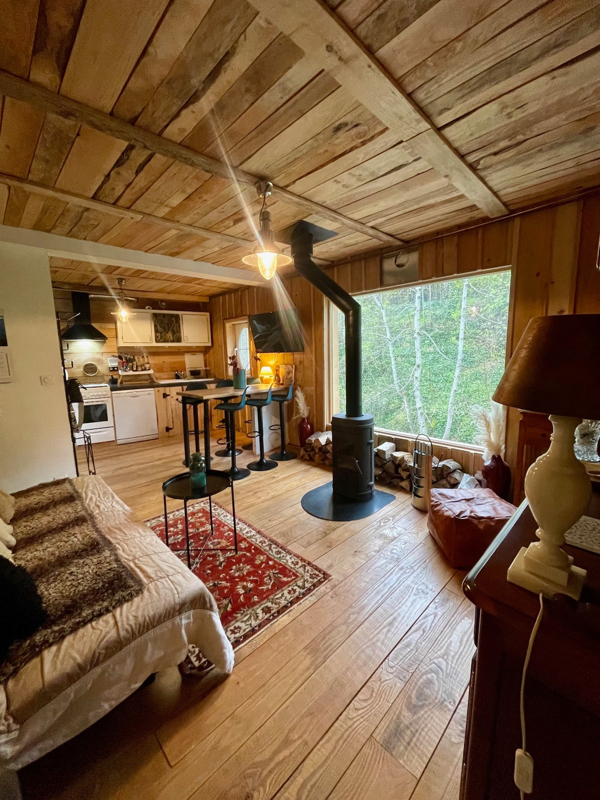 A cozy living area features wooden beams on the ceiling and warm wooden flooring. A small dining set with high chairs is positioned next to a window overlooking greenery. A wood stove stands prominently, accompanied by a patterned rug, creating a comfortable ambiance.