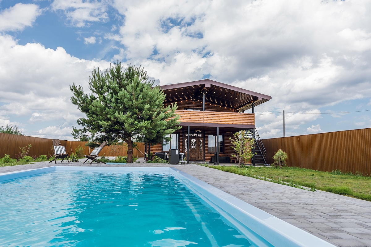 A sunny outdoor area features a clear swimming pool in the foreground. A wooden house with a large deck and multiple steps is set against a backdrop of partly cloudy skies. Lush greenery surrounds the property, complemented by lounge chairs positioned nearby.