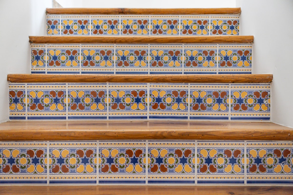 A set of stairs is adorned with colorful tiled risers featuring intricate floral patterns. The warm wooden steps contrast the vibrant tiles, creating a visually appealing ascent. Natural light highlights the textures and colors of the staircase.