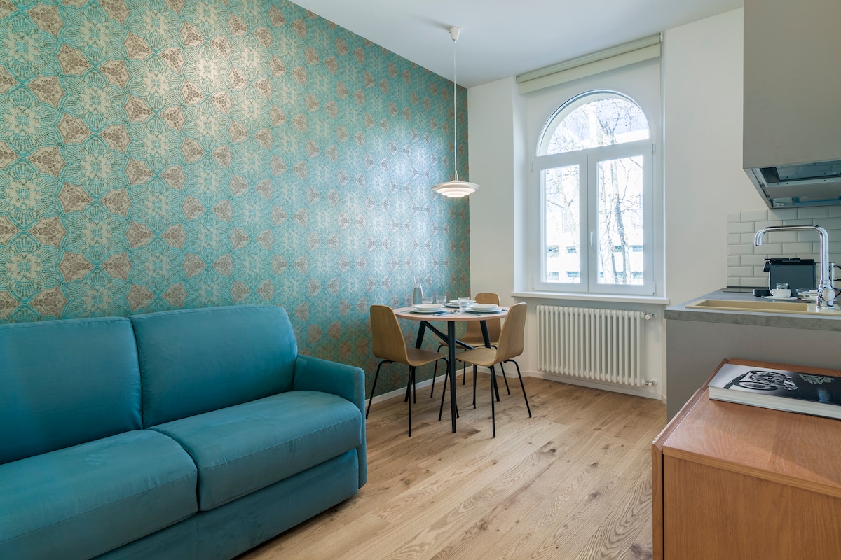 A welcoming living area features a blue sofa and a dining table with four chairs, set against a textured teal wallpaper. Natural light enters through a large arched window, illuminating the wooden flooring and modern kitchen area in the background.