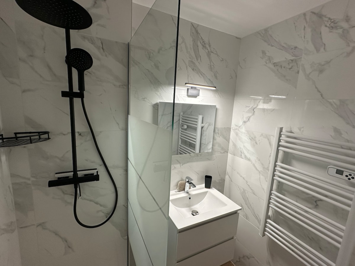 The modern shower room features a glass-enclosed shower equipped with a large overhead rain shower head. A wall-mounted mirror reflects soft lighting above a minimalist sink with storage underneath. White marble-like tiles create a clean and elegant aesthetic, complemented by a heated towel rail.