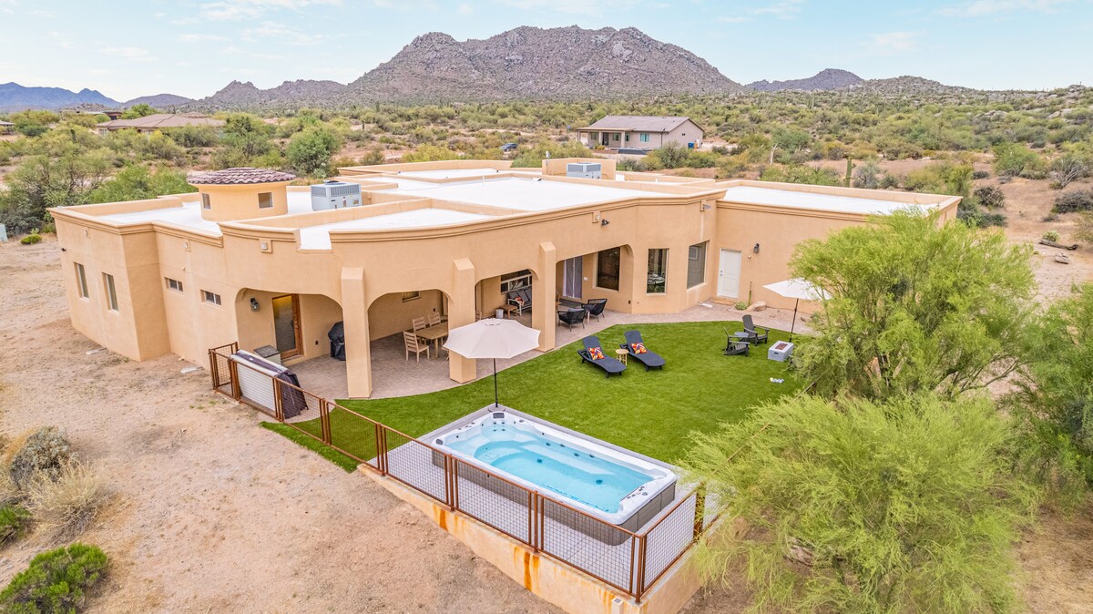 The spacious desert home is set against a backdrop of rolling hills. A well-maintained backyard features a hot tub, lounge chairs, and an umbrella for shade. The exterior showcases a blend of natural materials and large windows, creating a seamless connection with the surrounding landscape.