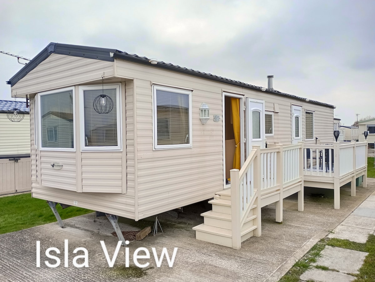 The exterior of the caravan presents a light-colored façade with multiple windows, allowing ample natural light. A deck area with steps provides access to the entrance. The surrounding grassy area features additional neighboring units in a seaside setting.