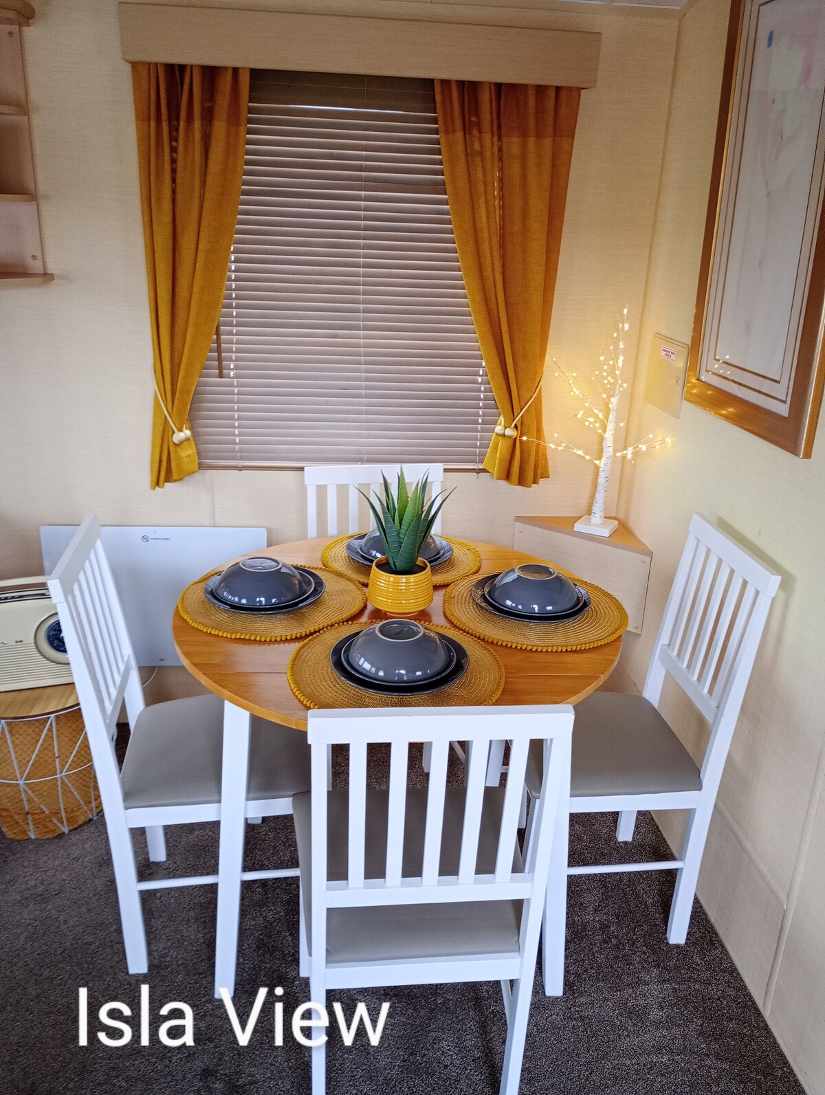 A round dining table is set with grey plates at each place setting. Four white chairs surround the table. A small potted plant and a decorative light feature sit at the center. Warm curtains frame the window, allowing soft light to enhance the space.