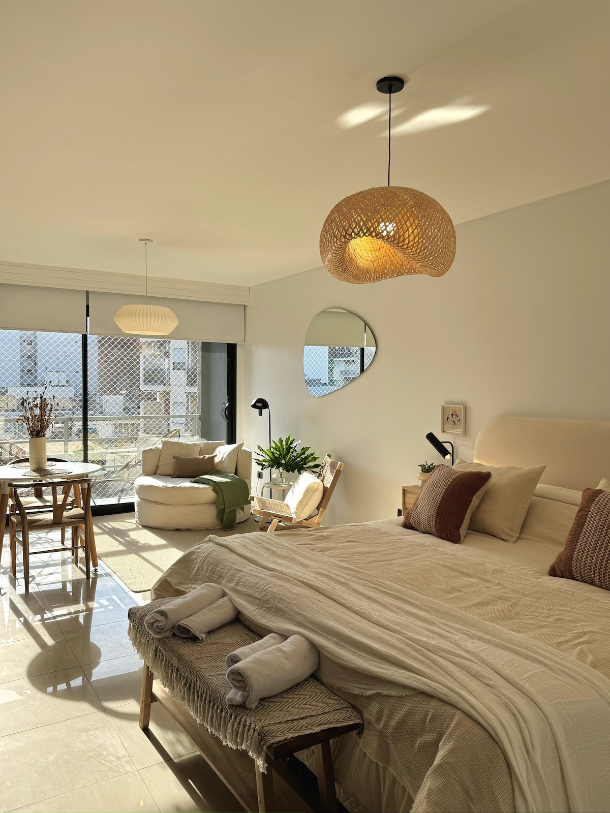 A bright studio is furnished with a spacious bed and decorative pillows. A small dining table with two chairs sits near a cozy lounge chair. Natural light is enhanced by light fixtures and a round mirror, creating a welcoming and functional environment.