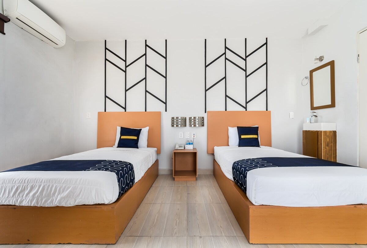 The room features two individual beds with simple wooden frames, accented by navy blue beddings. A small bedside table is positioned between the beds, with a lamp and essentials placed on it. Light-colored walls and wooden flooring contribute to an airy atmosphere.