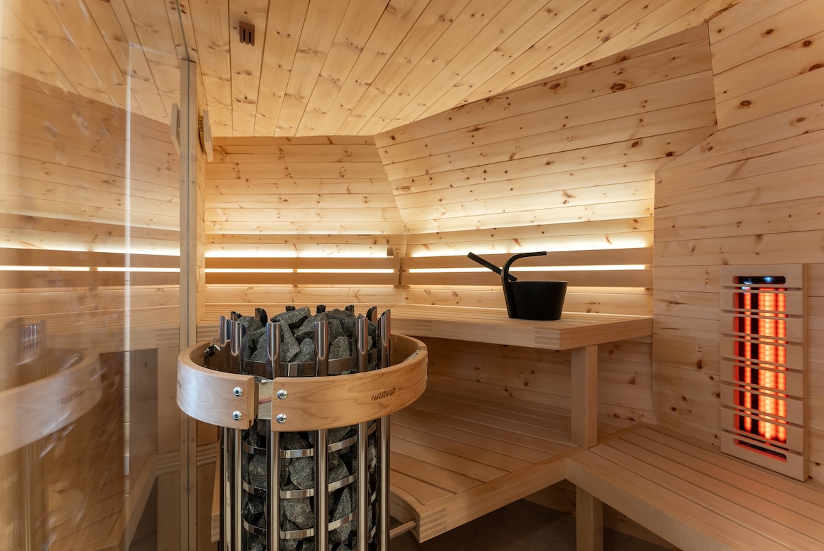 The sauna features warm wooden walls and benches, creating a soothing environment. A stone heater is centrally placed, surrounded by wood accents. An infrared panel provides additional warmth, with soft lighting enhancing the serene atmosphere, perfect for relaxation after outdoor activities.