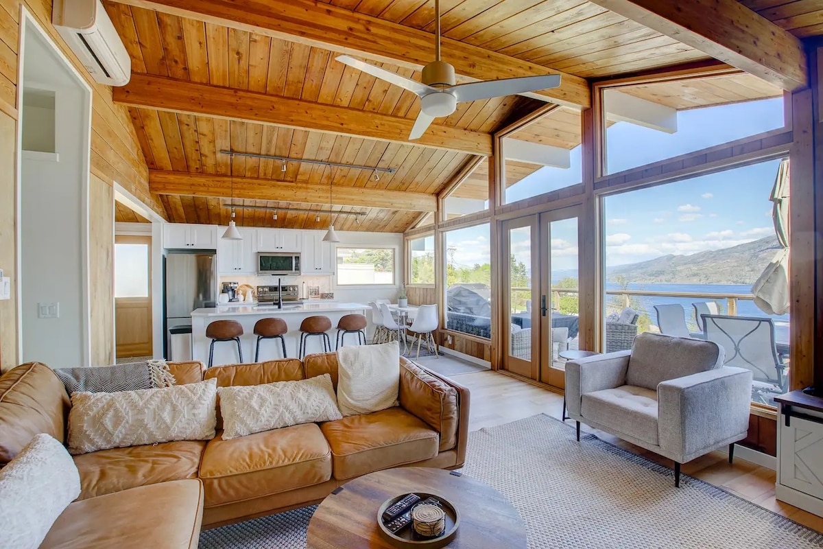 A spacious living area is characterized by wooden ceilings and large glass windows, providing views of the surrounding landscape. Comfortable seating includes a leather sofa and an armchair, with a circular coffee table at its center. The kitchen area features modern bar stools and a dining table in the background.