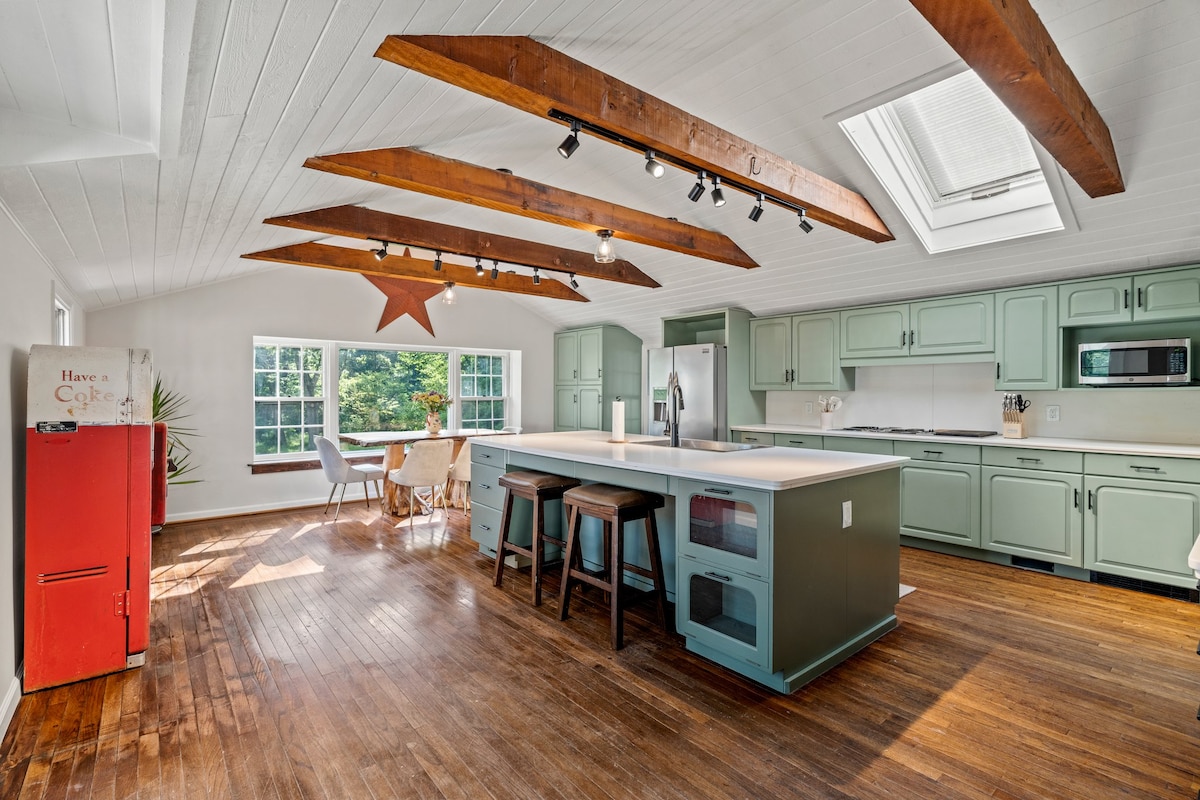 The kitchen features light green cabinets and a large island with seating for three. Skylights and windows allow natural light to fill the space, revealing rich wooden beams overhead. A red fridge adds a pop of color, enhancing the open and airy feel.