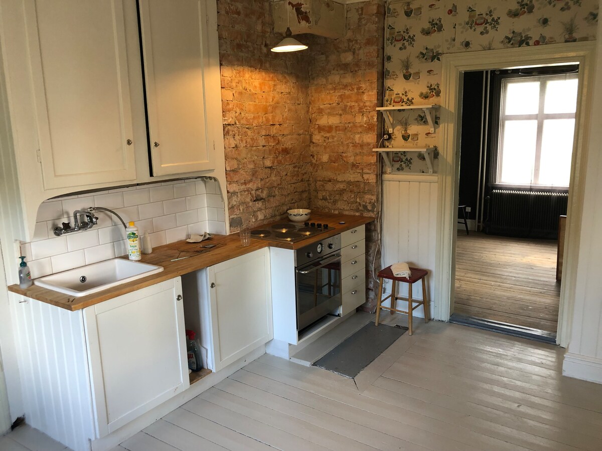 The kitchen area features white cabinetry paired with a wooden countertop and a modern stove. Exposed brick enhances the rustic charm, while light from the ceiling fixture illuminates the space. A small chair is positioned nearby, and wall-mounted shelving adds practicality.